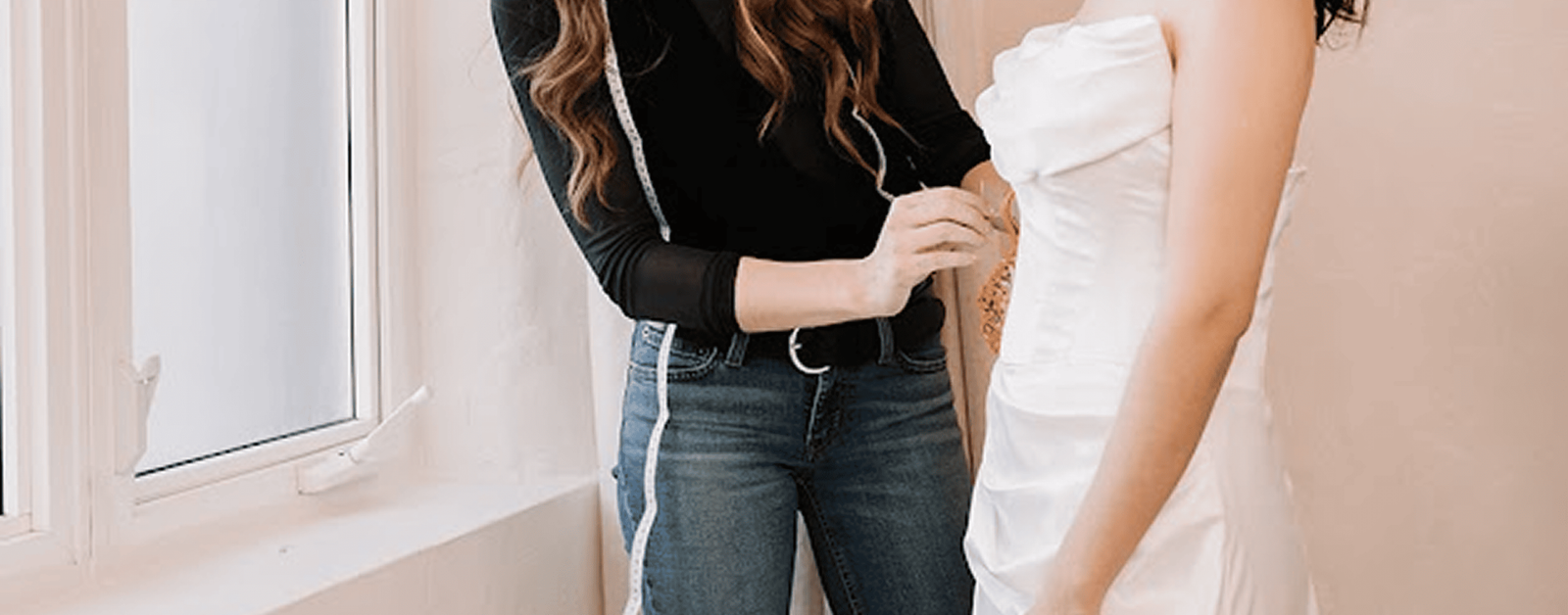 Tailor carefully adjusting the fit of a wedding dress on a bride standing by a bright window